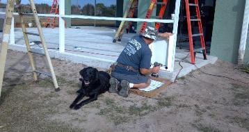 A technician kneels beside a black dog at an aluminum framing job site with ladders nearby.