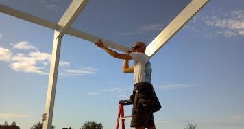 A technician on a ladder installs white aluminum roof framing against a blue sky.