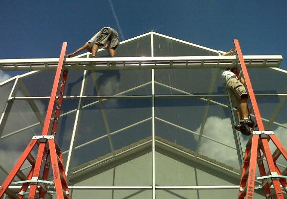 Two technicians on ladders install white aluminum gable-roof framing against a blue sky.